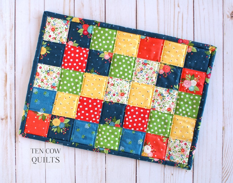 May include: A colourful patchwork quilt placemat with a variety of floral and geometric patterns in shades of red, yellow, green, blue, and white. The placemat is edged in a dark blue fabric and has the text "TEN COW QUILTS" in white lettering.