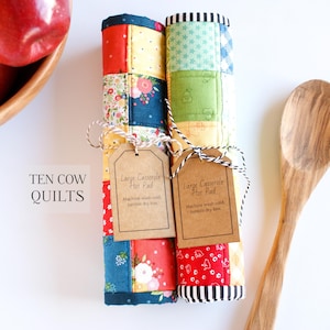 May include: Two colourful quilted casserole hot pads with brown tags that read "Large Casserole Hot Pad" and "Machine wash cold, tumble dry low". The hot pads are rolled up and tied with twine. The text "TEN COW QUILTS" is visible in the bottom left corner of the image.