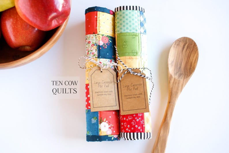 May include: Two colourful quilted casserole hot pads with brown tags that read "Large Casserole Hot Pad" and "Machine wash cold, tumble dry low". The hot pads are rolled up and tied with twine. The text "TEN COW QUILTS" is visible in the bottom left corner of the image.