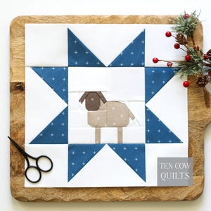 Könnte beinhalten: Ein quadratischer Stoff-Quiltblock mit einem zentralen, zusammengesetzten Schaf-Design in Braun und Beige, umgeben von einem blau-weißen Sternmuster mit Kreuzstich-Motiv. Der Text TEN COW QUILTS ist in der unteren rechten Ecke aufgedruckt.