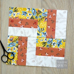 Könnte beinhalten: Ein Patchwork-Quiltblock mit floralen und geometrischen Mustern. Der Block zeigt gelbe, orangefarbene, weiße und blaue Stoffe. Eine schwarze Schere befindet sich in der unteren linken Ecke. Der Text "TEN COW QUILTS" befindet sich in der unteren rechten Ecke.