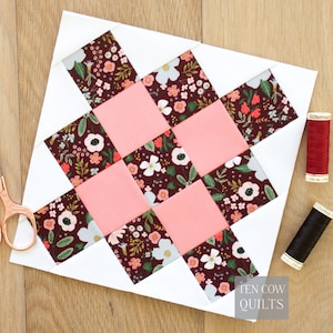 May include: A quilt block with a woven pattern of pink squares and floral fabric squares on a white background. The floral fabric has a dark red background with white and pink flowers. The image includes scissors and spools of thread.