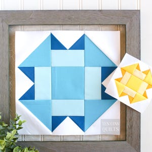 Op de afbeelding: Een ingelijst quiltblok met een geometrisch patroon in blauw en wit. Het blok is omgeven door een witte achtergrond en een grijs frame. De tekst "TEN COW QUILTS" is zichtbaar in de rechterbenedenhoek van het frame.