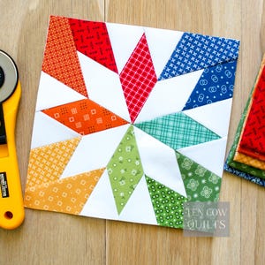Può includere: Un blocco di patchwork colorato con un motivo a stella. La stella è composta da tessuti fantasia in rosso, arancione, giallo, verde, blu e bianco. Sono visibili anche un taglierino rotante e quadrati di tessuto.