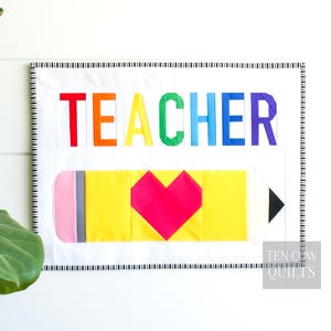 May include: A colorful quilt with the word "TEACHER" in rainbow hues above a yellow pencil with a pink eraser and a red heart. The quilt has a black and white striped border and is a teacher-themed item.