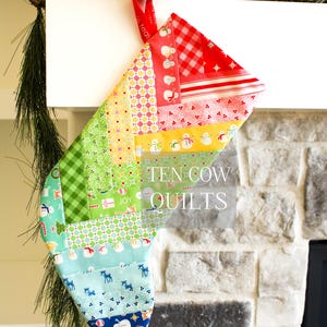 May include: A colorful patchwork Christmas stocking with a green, red, yellow, blue, and white pattern. The stocking is hanging on a white mantel and has the text "TEN COW QUILTS" on it.