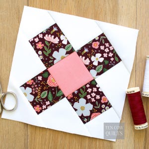 May include: A quilt block with a central pink square and four surrounding floral print rectangles on a white background. The floral print features pink, white, and blue flowers on a dark burgundy background. Scissors and spools of thread are visible.