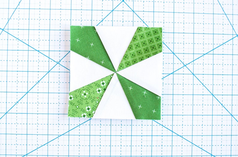 May include: A white fabric square with eight green fabric triangles sewn around the edges. The triangles have different patterns, including small white flowers, small white diamonds, and white crosses.