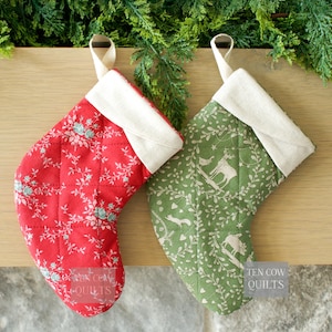 Peut inclure: Deux bas de Noël matelassés. L'un est rouge avec un motif floral, l'autre vert avec un motif de cerf et de forêt. Les deux ont un revers et une boucle de suspension crème. Ils sont présentés sur une surface en bois.