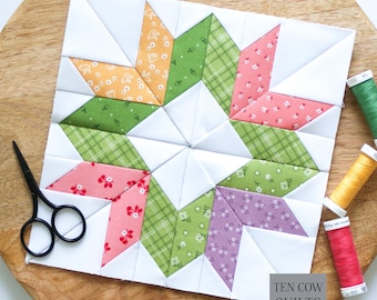May include: A square quilt block featuring a star pattern made of white, orange, green, pink, and purple floral and plaid fabric pieces. Black scissors are positioned on the left, and three spools of green, yellow, and pink thread are on the right.