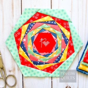 Może przedstawiać: Kolorowy, sześciokątny blok do pikowania z spiralnym wzorem. Blok zawiera czerwone, żółte, niebieskie i zielone tkaniny. Środek ma czerwony sześciokąt z białym haftowanym sercem i wzorem igły. Tekst "TEN COW QUILTS" znajduje się w prawym dolnym rogu.