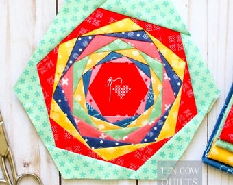 May include: A colorful, hexagonal quilt block with a spiral design. The block features red, yellow, blue, and green fabrics. The center has a red hexagon with a white embroidered heart and needle design. The words "TEN COW QUILTS" are in the bottom right corner.