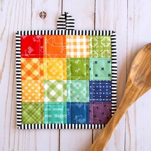 May include: A square quilted potholder featuring a patchwork grid of multicolored fabric squares in red, orange, yellow, green, blue, and purple with various patterns. It has a black and white striped border and a matching hanging loop.