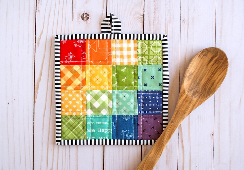 May include: A square quilted potholder featuring a patchwork grid of multicolored fabric squares in red, orange, yellow, green, blue, and purple with various patterns. It has a black and white striped border and a matching hanging loop.