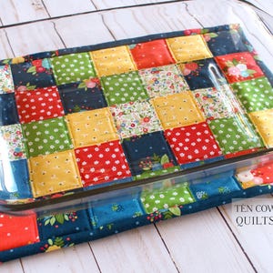 May include: A colourful patchwork quilted hot pad with a glass baking dish on top. The hot pad is made of squares of fabric in various colours and patterns, including red, blue, green, yellow, and white. The text "TEN COW QUILTS" is visible in the bottom right corner of the image.