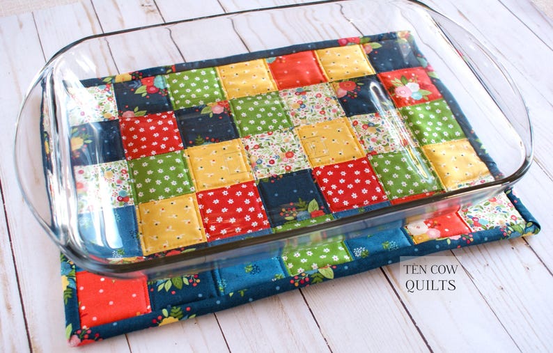 May include: A colourful patchwork quilted hot pad with a glass baking dish on top. The hot pad is made of squares of fabric in various colours and patterns, including red, blue, green, yellow, and white. The text "TEN COW QUILTS" is visible in the bottom right corner of the image.