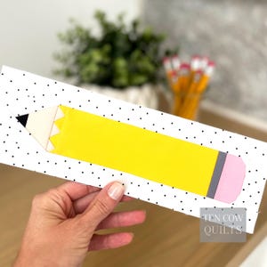 Pode incluir: Um desenho em forma de lápis amarelo com ponta preta e borracha rosa, sobre um fundo branco com bolinhas pretas. O desenho está sobre uma superfície branca. O texto "TEN COW QUILTS" é visível no canto inferior direito.