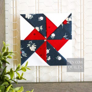 Pode incluir: Um bloco de colcha quadrado com um design de cata-vento. O bloco apresenta tecido floral branco, vermelho e azul marinho. O texto "TEN COW QUILTS" &eacute; vis&iacute;vel no canto inferior direito. O bloco &eacute; exibido em um fundo branco.