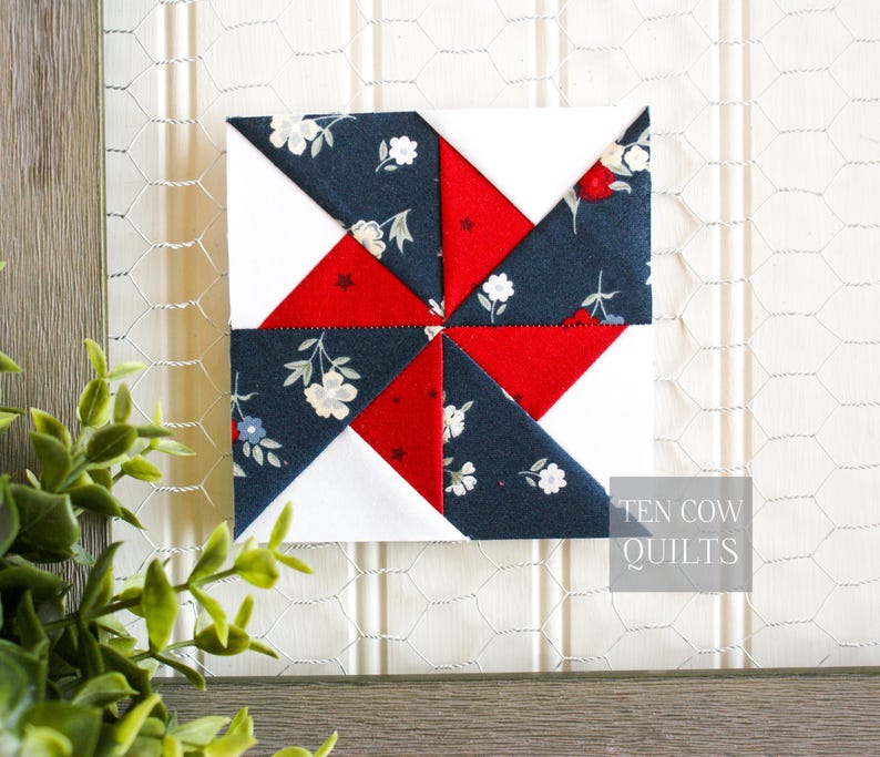 Pode incluir: Um bloco de colcha quadrado com um design de cata-vento. O bloco apresenta tecido floral branco, vermelho e azul marinho. O texto "TEN COW QUILTS" &eacute; vis&iacute;vel no canto inferior direito. O bloco &eacute; exibido em um fundo branco.