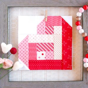 May include: A pink and red patchwork heart quilt block with a white background. The block is made up of various fabrics with different patterns, including polka dots, stripes, and hearts. The block is framed in a gray frame with a wire mesh background.