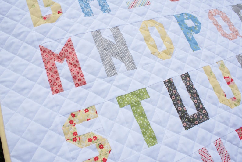 May include: A white quilt with a diamond pattern features a patchwork alphabet in various colors and patterns. The letters are made from different fabrics with floral and geometric designs.