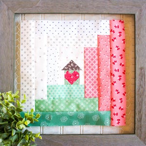 Könnte beinhalten: Ein gerahmter Patchwork-Quiltblock mit einem Hausdesign. Das Haus ist aus braunem, rotem und grünem Stoff gefertigt. Der Quiltblock ist von einem weißen Rahmen umgeben.