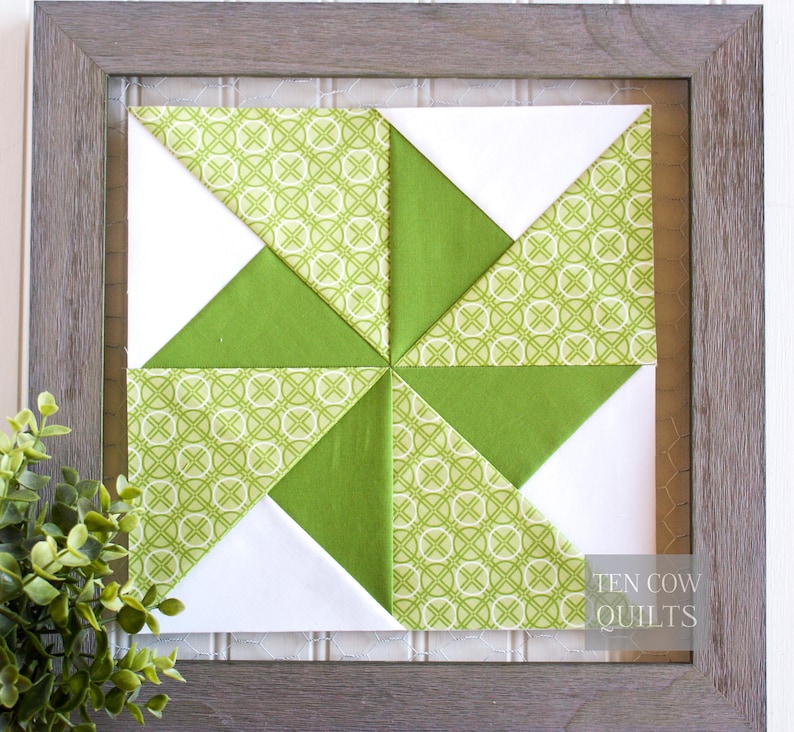 Pode incluir: Um bloco de colcha emoldurado com um design de cata-vento. A colcha apresenta tecido branco, verde liso e verde estampado. O tecido estampado tem um design geom&eacute;trico verde claro. A moldura &eacute; de cor cinza claro e o texto "TEN COW QUILTS" &eacute; vis&iacute;vel.