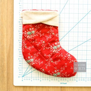 May include: A quilted Christmas stocking with a red floral pattern and a cream-coloured cuff. The stocking is diamond-stitched and features a delicate floral design in white and light green. The text "TEN COW QUILTS" is visible.