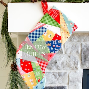 Puede incluir: Una media de Navidad de patchwork con un esquema de color rojo, blanco y azul. La media está hecha de varias telas con diferentes patrones, incluyendo cuadros, lunares y copos de nieve. La media está colgada en una repisa blanca con una guirnalda verde.