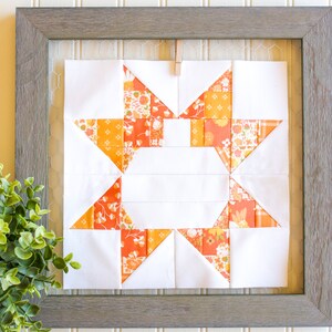 Pode incluir: Um bloco de colcha emoldurado com um design de estrela em tecido laranja e branco. A estrela é feita de quadrados e triângulos menores. O bloco de colcha está pendurado em uma parede branca.