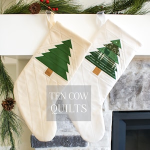 May include: Two white quilted Christmas stockings with green patchwork tree designs. The stockings are hanging from a white mantel above a fireplace. The text "TEN COW QUILTS" is printed in the center of the image.