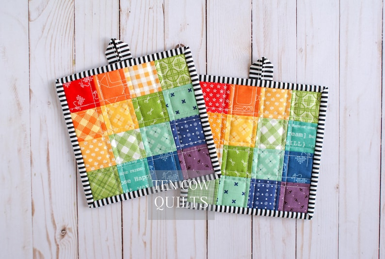 May include: Two square quilted potholders featuring a patchwork design of multicolored fabric squares in shades of red, orange, yellow, green, blue, and purple. Each potholder has a black and white striped border and hanging loop. Text reads TEN COW QUILTS.