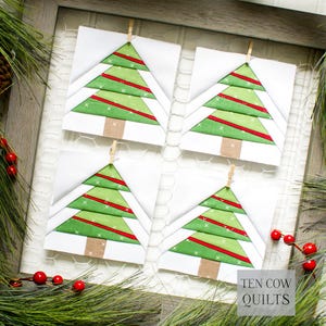 May include: Four Christmas tree quilt blocks, each featuring a green tree with red stripes and a brown trunk, are displayed. The blocks are clipped to a white background with small wooden clothespins. The image is framed with greenery and red berries.