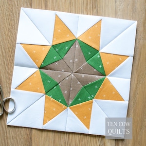 May include: A square quilt block featuring a star design. The star is composed of brown, green, and yellow triangles, with white cross-stitch details. The background is white. The text "TEN COW QUILTS" is in the lower right corner.