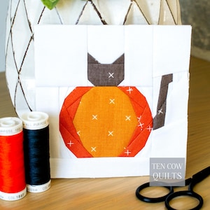 Puede incluir: Un bloque de patchwork blanco y naranja con un gato sentado sobre una calabaza. El gato es marrón y la calabaza es naranja. Sobre una superficie de madera, con carretes de hilo y tijeras. El texto "TEN COW QUILTS" es visible.