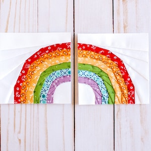 May include: Two fabric squares with a rainbow design sewn together. The rainbow is made of multiple fabric strips in red, orange, yellow, green, blue, and purple. The fabric squares are on a white wooden background.