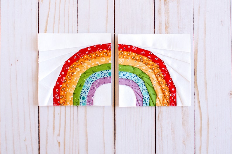 May include: Two fabric squares with a rainbow design sewn together. The rainbow is made of multiple fabric strips in red, orange, yellow, green, blue, and purple. The fabric squares are on a white wooden background.