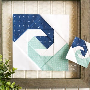 May include: A framed quilt block with a spiral design in blue and green fabrics. The block is displayed against a white background with a wire mesh behind it. The text "TEN COW QUILTS" is visible in the lower right corner.