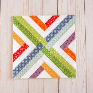 K&ouml;nnte beinhalten: Ein Patchwork-Quiltblock mit wei&szlig;em Hintergrund und diagonalen Stoffstreifen in verschiedenen Farben, darunter Rot, Orange, Gelb, Gr&uuml;n, Blau und Lila.