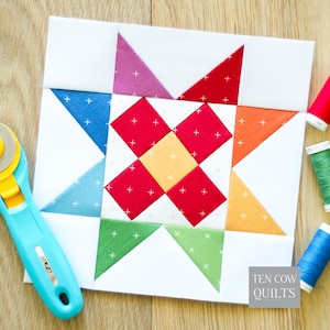 May include: A square quilt block featuring a central yellow square surrounded by four red squares, with outer triangles in blue, pink, red, orange, yellow, and green, all with a white plus-sign pattern. A rotary cutter and three spools of thread are nearby.