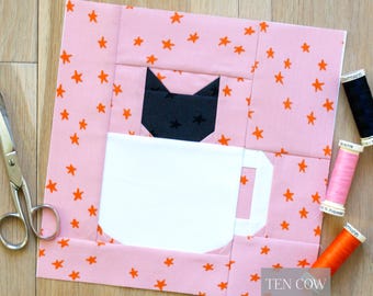 May include: A pink and orange star patterned quilt block featuring a black cat head above a white coffee mug. The block is surrounded by scissors and spools of thread. The text "TEN COW QUILTS" is in the bottom right corner.