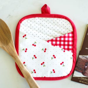 May include: A square, quilted pot holder with a red border and hanging loop. The pot holder features three fabric sections: white with black crosses, white with red cherries, and red and white gingham. A wooden spoon is next to the pot holder.