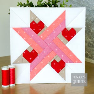 Puede incluir: Un bloque de edredón hecho a mano con un diseño de estrella y cuatro formas de corazón rojas. La estrella está hecha de tiras de tela rosa, melocotón y beige, con detalles de punto de cruz blancos. El texto "TEN COW QUILTS" es visible. Dos carretes de hilo rojo están en primer plano.