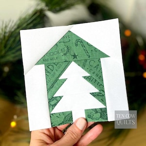 May include: A handmade Christmas tree quilt block. The block features a white tree shape inside a green house shape. The green fabric has a pattern of candy canes and text that reads "Cookies and Milk". The block is on a white background.