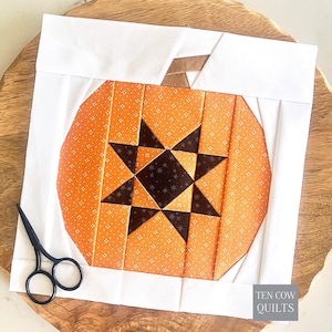 May include: A square fabric quilt block featuring a pumpkin design constructed from orange fabric with a white star pattern, a dark brown star center, and a brown stem. The block is framed by white fabric borders and includes a pair of black scissors.