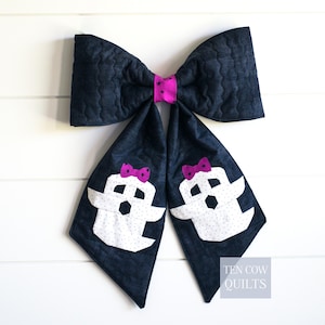 May include: A large quilted hair bow made of dark navy fabric with a wavy texture. The center knot is bright pink with black stars. Two long tails feature white ghost appliqués with tiny black dots and small pink bows on their heads. Text reads TEN COW QUILTS.