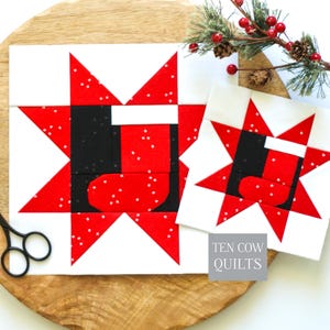May include: Two square quilt blocks featuring a red and white star pattern with a central black and red stocking design. The fabric has a subtle white speckled print. A grey square label with the text TEN COW QUILTS is visible on the smaller block.