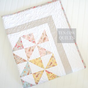 May include: A white quilt with a patchwork design featuring pink, yellow, and blue floral fabric. The quilt has a light brown border and is quilted with a wavy pattern. The text "TEN COW QUILTS" is printed in the top right corner.