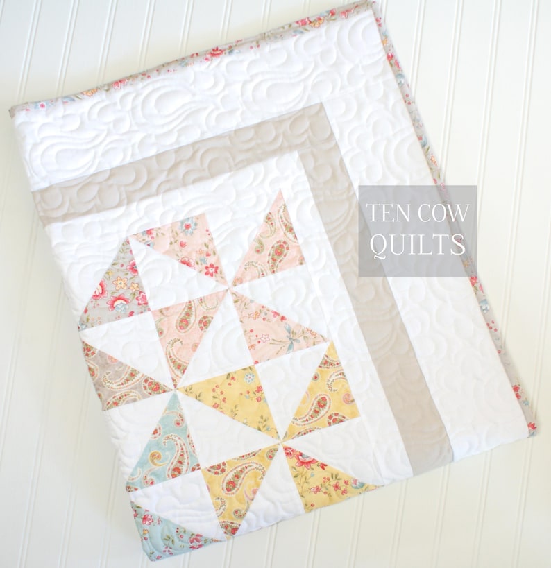 May include: A white quilt with a patchwork design featuring pink, yellow, and blue floral fabric. The quilt has a light brown border and is quilted with a wavy pattern. The text "TEN COW QUILTS" is printed in the top right corner.
