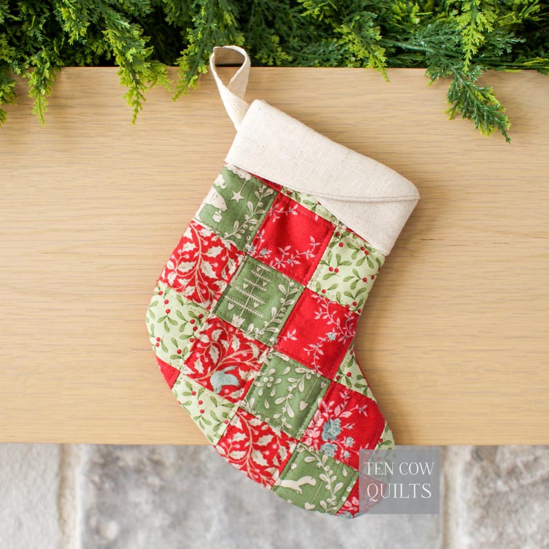 May include: A patchwork Christmas stocking in red and green with floral and festive patterns. It has a cream-coloured cuff and hanging loop. The text "TEN COW QUILTS" is visible. The stocking is made of quilted fabric.